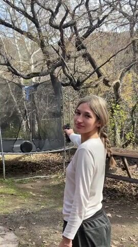A young woman with blonde hair in a braid, wearing a white long-sleeved shirt and dark sweatpants, stands in a backyard setting and points a black aerosol can towards a trampoline. The trampoline is enclosed with a black net and has a black disc on its surface. The woman sprays the can, and a mist appears. She then turns and smiles towards the camera. Trees with bare branches and some greenery are visible in the background, suggesting a cooler season. Sunlight filters through the trees. A picnic table is visible on the right side of the frame.
