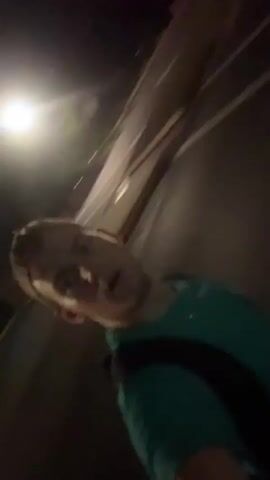 A man wearing a teal shirt and a backpack films himself from a low angle at night. He appears to be on a street with streetlights and traffic signals. The man is looking towards the camera. The camera is tilted, giving a dynamic perspective. The background is blurred, suggesting movement.