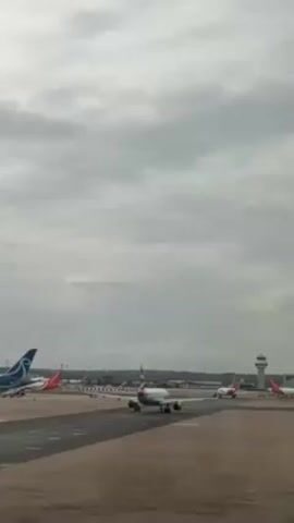 The video shows an airport runway on a cloudy day. Several airplanes are visible, including one with a red and white livery that is closer to the camera and appears to be taxiing. Other planes are parked in the background, with one featuring a blue tail. An airport control tower stands to the right of the frame. The ground is dark and wet, suggesting recent rain. The overall scene depicts typical airport activity under overcast conditions.