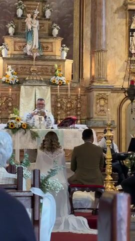 The video shows a wedding ceremony in a church. A priest stands behind an altar decorated with sunflowers and lilies. He is holding a chalice. In front of the altar, a bride and groom are kneeling. The bride is wearing a white dress with a long veil, and the groom is wearing a green suit. Behind them, wedding guests sit in pews. An ornate altar is visible in the background, featuring a statue of Mary and baby Jesus.  A musician sits off to the side, playing an instrument.