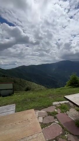 The video pans across a mountainous landscape under a partly cloudy sky. In the foreground, a wooden picnic table is visible. To the left of the table, there is a grassy area with a white sign and some seating. As the camera pans, the view shifts to a green mountain range with scattered trees. A Bulgarian flag on a flagpole stands prominently on a hilltop, with a further mountain peak in the background. The environment appears to be a scenic overlook or a hiking destination.