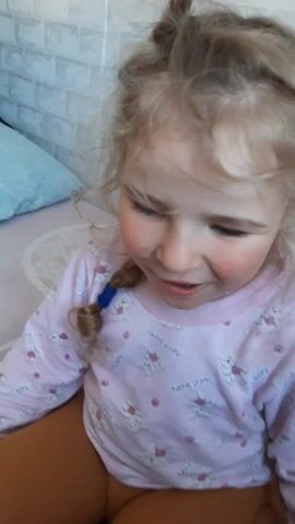 The video shows a young girl with blonde hair and blue eyes. She is wearing a pink long-sleeved shirt with teddy bear designs and brown pants. Her hair is partially braided. The girl smiles at the camera. The background includes a white brick-patterned wall and part of a bed with a patterned blanket.