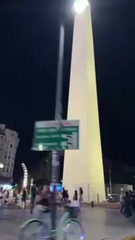 The video shows a tall, white obelisk at night, possibly a monument or landmark. It stands in a plaza or public space, with people walking around it. There are buildings visible in the background, lit up against the dark sky. The camera appears to be moving slightly. Traffic lights are visible nearby, adding to the urban atmosphere.