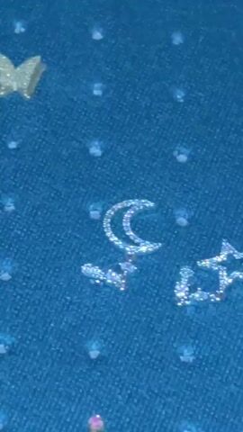 This is a close-up video of various glittering objects and jewelry. The initial frames show a blue surface with what appear to be scattered glittery shapes resembling moons and stars. These are followed by frames that reveal rose gold glittery embellishments, possibly flowers or abstract designs, on a dark textured surface, likely fabric. The video then transitions to show a collection of rings stored in a dark green velvet-lined jewelry box. The rings are varied in style, with some featuring bands encrusted with small stones and others showcasing larger gemstones, including a prominent square-cut stone with rainbow reflections. The overall impression is one of shiny, decorative items, with a focus on sparkle and intricate detail. No people, specific time of day, weather, or location cues beyond Gudauta, Abkhazia are evident. No visible text is present.