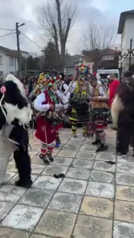 In a medium shot, a group of elaborately costumed men dance on a paved surface surrounded by onlookers. The dancers wear colorful outfits consisting of white shirts, brightly colored vests and skirts, and masks adorned with feathers and other decorations. Bells are attached to their costumes, creating a cacophony of sound as they move. The background features trees, buildings, and a crowd of people observing the spectacle. The overall atmosphere suggests a festive or ceremonial occasion taking place in an outdoor setting.