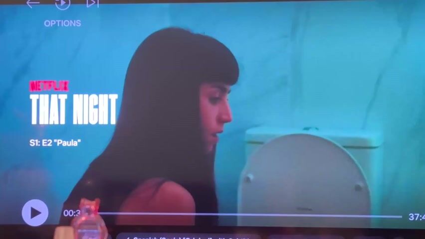 The still shot shows a TV screen streaming the Netflix series "That Night," specifically Season 1, Episode 2 titled "Paula." The image primarily features a woman with dark hair and bangs, shot in profile from the shoulders up. She appears to be in a bathroom, as a toilet is visible in the background. There's a play button in the lower left corner of the screen, indicating that the show is paused. Time stamps "00:37" and "37:44" are visible, along with "Spanish (Spain) [Original] with Subtitles" and "English [Audio Description]". A small, decorative perfume bottle is partially visible at the bottom left edge of the screen. The overall color tone of the image is bluish.
