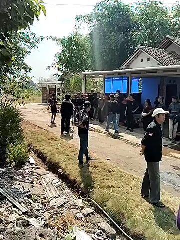 A group of approximately 20-25 people, mostly men wearing dark clothing, are gathered outdoors on a dirt road in front of a building with a porch. Some individuals are standing and talking, while others appear to be watching something or someone. There are several blue screens visible on the wall of the building, possibly monitors or displays. The environment appears to be rural, with vegetation and trees in the background. The ground is uneven and shows signs of construction or debris in the foreground. The lighting suggests it is daytime.