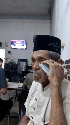 A man of advanced age is on a phone call. He wears a black peci hat with green embroidery and a light-colored shirt with embroidered details. He holds a silver-colored smartphone to his right ear. A red ring is visible on his right hand. He has gray hair and a mustache. The setting appears to be an indoor area, possibly a cafe or waiting room. In the background, a television displays an image, a water dispenser, and other individuals seated at a table. One person is partially visible to the left of the main subject. The room has tiled walls, and there is a fan nearby.