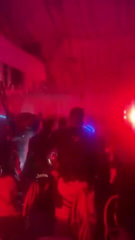 The video shows a group of people in a dimly lit room with red lighting. The room appears to be covered by a white fabric ceiling. Some individuals are wearing dark clothing, and one person has what appears to be blue lights on their back. A bright red light with a yellow center shines into the camera.