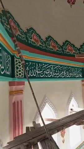 The video shows an interior view of what appears to be a religious building, likely a mosque. Above the viewer's vantage point, there is Islamic calligraphy inscribed on a green band along the wall.  This band is bordered by further decorative painting, including orange and green floral-like motifs. Beneath the calligraphy band, there are arched windows and decorative pink pillars. A wooden structure, presumably scaffolding, is visible in the foreground obscuring parts of the view. A sound that seems to be a call to prayer is audible.