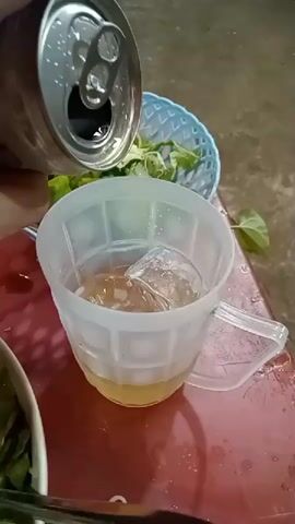The video begins with a close-up of someone pouring a light-colored liquid from a silver can into a white plastic mug filled with ice. Beside the mug are a small blue basket with green leaves and a white bowl with other food items. The scene then shifts to show a red table with yellow plastic chairs, indicating an outdoor eating area, possibly a restaurant or food stall. The camera pans across the space, revealing other red tables and yellow chairs occupied by people eating and using laptops. Overhead, the area is covered by a tarp or awning, and lights illuminate the scene, suggesting it is nighttime. The ground appears to be wet, likely from rain.