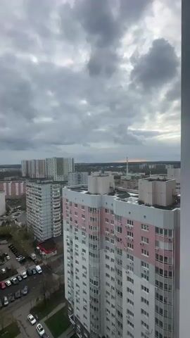 The video captures a wide view of an urban landscape in Gavrilkovo, Russia, from a high vantage point, likely a balcony or window of a tall building. The camera slowly pans downwards, revealing a bustling cityscape under a cloudy, overcast sky.

In the distance, numerous multi-story apartment buildings of varying architectural styles and colors dominate the skyline, some appearing newer with colorful facades (green, yellow, and blue elements) and others older, in shades of grey and beige. As the camera lowers, the view reveals the immediate vicinity below. A large residential complex with a prominent pink and white building is in the foreground on the right, and another large beige building with multiple windows is to the left.

Below, several parking lots are filled with parked cars of various models and colors. A busy road is visible, with vehicles driving and parked along the side. A yellow car, possibly an ambulance, is seen moving or parked near a small green area. A playground with colorful equipment is also visible in the courtyard between buildings. The overall impression is one of a dense, active residential area.

The sky is heavily overcast with dark grey clouds, but a sliver of lighter sky and a hint of orange light can be seen on the distant horizon, suggesting either early morning or late afternoon. The sounds of traffic, including a car horn, are audible throughout the video, emphasizing the urban setting.