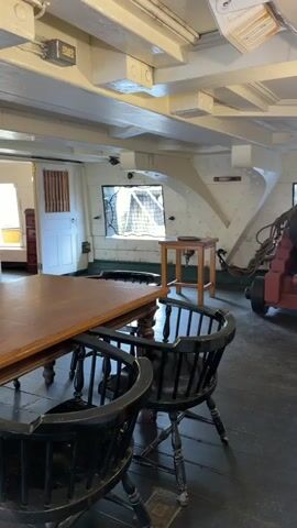 The video shows a room with a large wooden table and chairs. There are double doors in the background that open to another room. A fire extinguisher is visible on the left side of the table. A dresser with a mirror is also in the room. The room appears to be on a ship, with wooden beams in the ceiling and portholes. The lighting is dim.