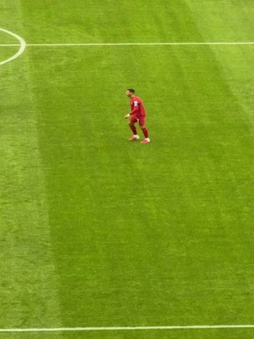 The video shows a soccer player in a red uniform with pink shoes and white socks walking across a bright green soccer field. The player is facing away from the camera and appears to be looking off to the side. The player's number 7 is visible on the back of their jersey. Another player in a light blue uniform is partially visible on the left side of the frame. The field has white markings visible in the upper left corner. The lighting suggests it is daytime.