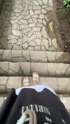 The video begins with a first-person view of a person's feet wearing white sandals as they walk down a stone staircase. The camera then pans up to show a stone path winding through a green, wooded area. A metal railing lines the left side of the path, and trees and foliage create a canopy overhead. The path leads towards a building visible in the distance.
