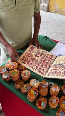 A person in a light green shirt is showing a book filled with pages covered with illustrations in color, laid out in grids, on a table covered with a green and red cloth. There are many round, flat, painted leather charms stacked and scattered on the table. The book is open and the person’s left hand holds open one page, as they point to an image on the other page. A black smartphone is on the table as well. The table is set outside in what seems to be a shaded area, with a yellow painted brick wall in the background.
