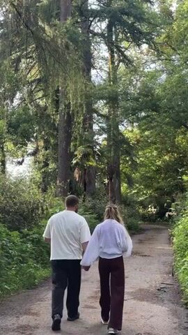 The video shows a man and a woman walking away from the viewer on a path in a forest. The man is wearing a white t-shirt and black pants. The woman is wearing a white shirt and dark brown pants. They are holding hands as they walk. The path is surrounded by tall trees and lush green vegetation. The lighting is bright and natural, suggesting it is daytime.