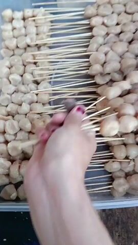 The video shows a close-up, overhead view of a plastic container filled with numerous small, light-colored meatballs skewered on thin wooden sticks. The meatballs are arranged in neat rows within the container. A hand, with fingernails painted purple and pink, is shown holding a cluster of these skewers and is in the process of either arranging them or selecting them from the container. The background is slightly blurred, but appears to be a rustic, possibly outdoor or kitchen setting with wooden surfaces visible. The lighting is natural and appears to be daylight.