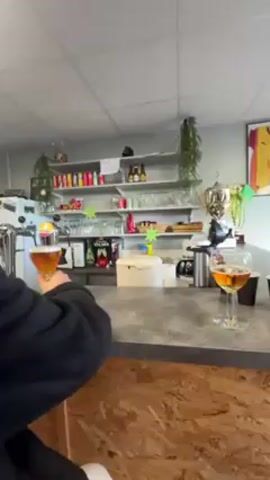 The video shows two people standing at a bar counter. The person in the foreground has curly hair and is wearing a black jacket. They are holding a glass of beer and looking at it. Another glass of beer sits on the bar counter next to them. The bar area has shelves stocked with various drinks and glassware. A small refrigerator, coffee maker, and some decorations can also be seen. The bar counter itself has a textured surface with a wood-like design on the front.