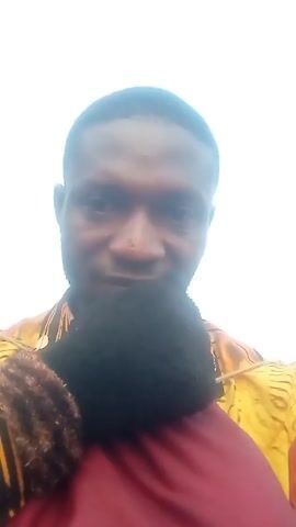 A man is looking at the camera. He has short dark hair and a slight beard. He is wearing a yellow and brown patterned shirt. He is holding a child with dark curly hair, who is wearing a maroon shirt. The background is a blurry white sky.