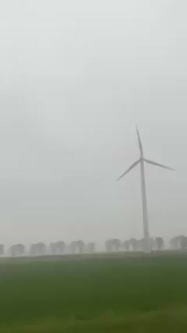 The video shows a field of green grass under a foggy, overcast sky. A line of thin trees runs along the horizon, and a tall wind turbine stands at the edge of the field. A cyclist rides across the field from left to right, passing in front of the turbine. Electronic music plays throughout the video.