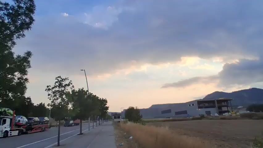 The video shows a wide shot of a rural landscape during sunset. The sun is low in the sky, partially obscured by clouds, casting a warm glow on the scene. In the foreground, there is a field of dry grass. In the middle ground, there is a road with a sidewalk and trees lining it. Several trucks are parked along the road. In the background, there are mountains and several industrial-looking buildings under construction. The sky is filled with dramatic, dark clouds that are illuminated by the setting sun.