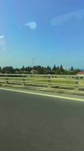The video shows a view from a moving vehicle, looking out a window. The perspective is from the side, capturing a landscape that includes a highway or road in the foreground. Beyond the road, there is a barrier with a railing, and further back, there are trees, houses with orange roofs, and a clear blue sky. The sun appears to be shining, creating bright conditions. The movement of the vehicle suggests a journey is in progress.