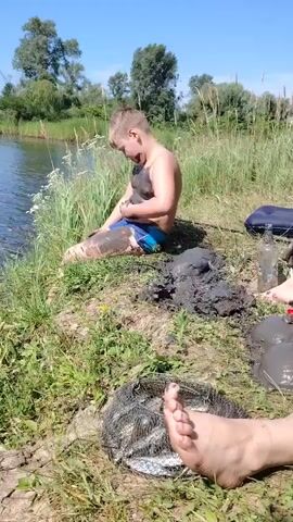 A boy covered in mud sits by the edge of a body of water. He is wearing blue swim trunks and his arms and legs are coated in dark mud. He is holding a muddy object in his hands and appears to be playing with it. Beside him, on the grassy bank, is a pile of mud, a mesh bag filled with fish, a partially inflated blue air mattress, and a clear glass bottle. The sun is shining, and the sky is clear blue, with trees visible in the background. The camera focuses on the boy and the surrounding area.