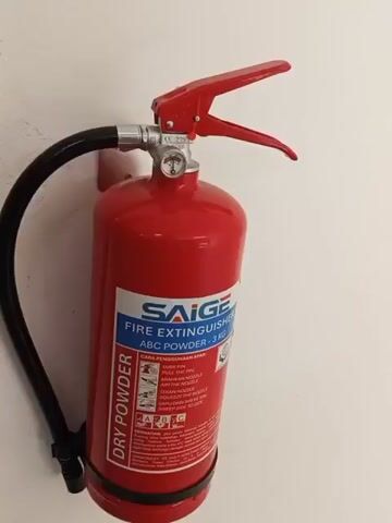 A close-up, eye-level, medium shot shows a red ABC dry powder fire extinguisher mounted on a white wall. The extinguisher is oriented vertically and is angled slightly to the right. The black hose and nozzle are visible to the left of the cylinder. The label on the extinguisher is legible and displays the brand name "SAIGE", "FIRE EXTINGUISHER", "ABC POWDER - 3 KG", and instructions for use in both English and another language, possibly Malay. The instructions include pictograms illustrating the PASS method: Pull the pin, Aim the nozzle, Squeeze the handle, and Sweep side to side. The gauge on top of the extinguisher indicates the pressure is within the green zone. The wall appears to be made of a textured material, possibly painted concrete or stucco. The lighting is even, suggesting an indoor environment. No people or other objects are visible in the frame.