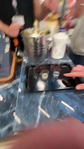 The video shows a person’s perspective as they take a shot from a tray being offered by someone. On the blue marble-patterned surface of a table, a tray with several small shot glasses, some with red liquid and some with clear liquid, is visible. A silver ice bucket with a bottle inside is also on the table. The person takes a shot glass filled with clear liquid from the tray. Two people are partially visible around the table. One is holding a white bottle. The other is wearing a black shirt and serving the shots. The environment appears to be an indoor event or possibly a retail setting, as indicated by other items on the table and background activity.