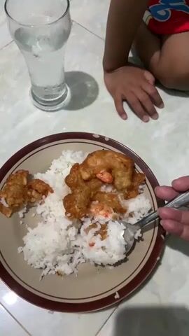 The video opens with a close-up of a plate of white rice and fried food, possibly shrimp, with a glass of ice water in the background. A hand holds a spoon with rice and food, bringing it towards the camera. The scene then shifts to a young child with dark hair and wearing a yellow t-shirt with colorful graphics, sitting on the floor. The child's mouth is open as a spoonful of rice is being offered. The child then takes the spoonful of rice into their mouth. The background shows a blurry interior with toys visible. The lighting is dim.