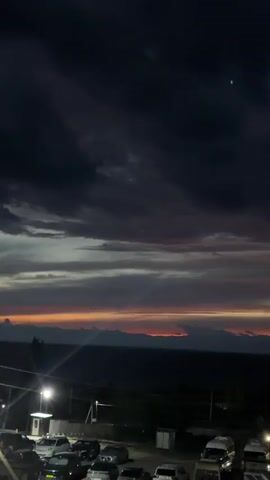 This is a vertical, low-angle video clip taken at dusk in Kadji-Sai 2, Kyrgyzstan. The scene is dominated by a dramatic sky filled with dark, ominous clouds that transition to streaks of orange and yellow near the horizon. This indicates either a sunset or an impending storm.

In the foreground, there is a dark, shadowy area that appears to be a parking lot or an open area with several vehicles. The vehicles are largely obscured by darkness, but some appear to be cars, vans, and possibly trucks. A single streetlamp on the left casts a pool of light on the ground. A few structures, possibly buildings or storage units, are visible in the lower part of the frame, some illuminated by artificial lights.

The middle ground is occupied by a body of water, likely a lake or sea, stretching across the frame. Beyond the water, a range of mountains is silhouetted against the colorful twilight sky. Their peaks are indistinct due to the low light conditions.

The overall mood of the video is somewhat somber and dramatic, owing to the dark, cloudy sky and the fading light. The presence of vehicles suggests a populated area or a place of transit. The natural elements – the stormy sky, the water, and the mountains – create a sense of scale and grandeur. There is no visible text in the video.