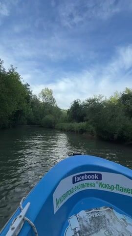 The video captures a boat ride on a river surrounded by lush greenery under a partly cloudy sky. The camera is initially focused on the front of the blue boat. A Facebook sticker and the text "Луковитска екопътека "Искър-Панега"" are visible on the side of the boat. The view then shifts to show a young woman smiling at the camera. Subsequently, the video reveals the other occupants of the boat, including a woman wearing sunglasses and an embroidered shirt, another young woman in a black tank top, and a man in a green shirt operating the motor. The atmosphere appears relaxed and joyful as the boat navigates the river.