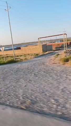 The video shows a view from a moving vehicle, likely a car or bus. The landscape is rural or semi-rural, with a clear blue sky and bright sunlight. In the background, there are structures that appear to be industrial or agricultural buildings, possibly greenhouses or storage facilities, and some rows of solar panels. A dirt road or path is visible in the foreground, with gravel and dry vegetation. A tall pole with a white triangular sign is also visible to the right. The overall impression is a sunny day in a sparsely populated area.