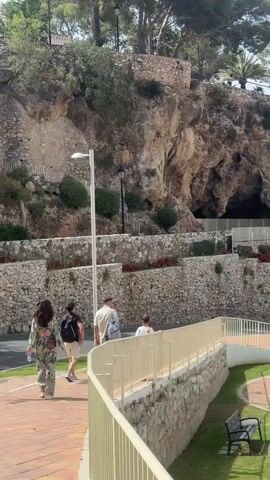 The video shows an outdoor scene with a rocky cliff face, terraced stone walls, and greenery. Several people are walking on a paved path in the foreground. In the background, there is a village with white buildings situated on a hill overlooking the sea. The sky is partly cloudy. The scene appears to be a tourist destination.