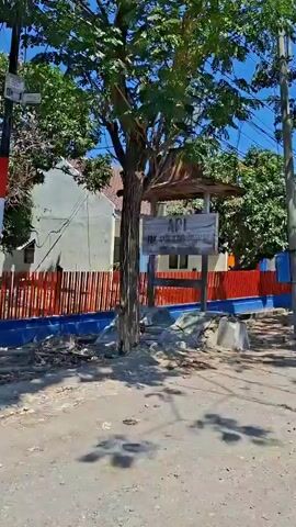 The video shows a moving shot of a residential or institutional area on a sunny day. The perspective is at street level, looking towards a building with a bright red and blue fence. Trees with green foliage are visible, some casting shadows on the ground. Electrical wires and poles are present, indicating urban infrastructure. Indonesian flags are displayed on poles along the fence, suggesting a national holiday or celebration. The overall scene is one of an outdoor environment with clear blue skies.