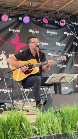 A man is sitting on a folding chair and playing an acoustic guitar. He is also singing into a microphone. He is wearing a black short-sleeved shirt, dark pants, and glasses. The guitar is light brown. The microphone is silver and attached to a stand. There is a music stand in front of him with sheet music on it. Behind him is a black backdrop with the word "BAKGARDEN" printed repeatedly in white. There are pink spotlights above the backdrop. In the foreground, there is green grass or foliage. The lighting is bright, and the scene appears to be outdoors, possibly on a stage. The man is looking towards the audience, and his mouth is open as if he is singing. He is strumming the guitar with his right hand and fingering chords with his left.