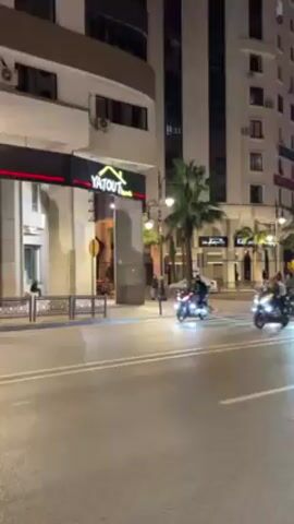 At night, three motorcycles, each carrying two people, ride on a paved road in an urban setting. The road is lined with streetlights, palm trees and buildings. The buildings have commercial signage, including the name "BATOUT." The people on the motorcycles wear dark jackets and helmets. The motorcycles have storage boxes attached to the back. There is a car behind the motorcycles. The motorcycles move from right to left in the frame.