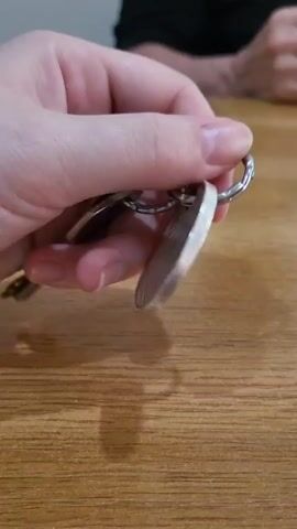 The video shows a person's hand holding a keychain with a coin attached. The coin is circular and appears to be made of silver. On one side of the coin, there is an image of a person and text that reads "THE STEADFAST TIN SOLDIER". On the other side, there is an image of a woman's profile and text that reads "FIJI" and "ONE DOLLAR". The keychain also has two keys attached. The background is a wooden table.