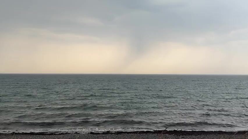 The video shows a wide, static shot of a choppy, grey ocean under an overcast sky. In the distance, a funnel cloud, potentially a waterspout, extends from the clouds to the water, appearing as a cone-shaped vortex. The foreground consists of a pebble beach with some dry grass or reeds visible at the very bottom of the frame. The lighting is dim, suggesting an impending storm or a generally cloudy day. There are no people or discernible objects in the water or on the beach.