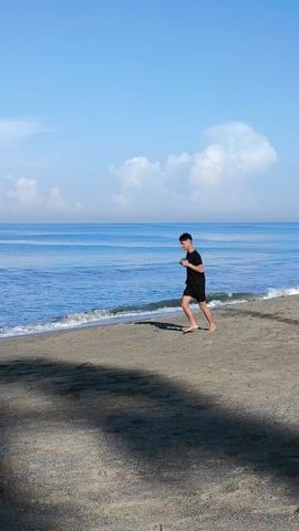 A young man jogs barefoot along a beach, wearing a black t-shirt and shorts. He briefly pauses to adjust his hair. The beach is sandy, and the ocean is calm, with a few people swimming and surfing in the distance. The sky is blue with scattered white clouds. The beach has wet sand on the shoreline, with a dark shadowed area in the foreground.