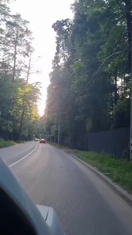 The video shows a POV shot from inside a vehicle traveling down a paved road lined with trees and a dark fence. Sunlight filters through the trees, creating a dappled effect on the road. Several cars are visible ahead, including a red one. There are also some streetlights and bus stop signs along the side of the road. The scene appears to be a suburban or rural area.