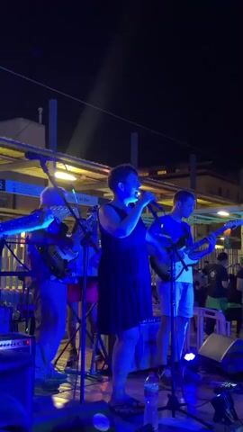 A band is performing on a stage outdoors at night. The band consists of four people, three men and one woman. The woman is in the center of the stage singing into a microphone. She is wearing a dark dress. To her left, two men are playing guitars. To her right, a third man is playing a guitar. The stage is lit with blue and purple lights. There are speakers and other musical equipment on the stage. In the background, there is a building with lights on. There are also people in the background watching the performance. The overall atmosphere appears to be festive.
