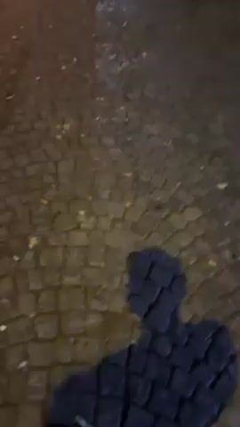 The video shows a street scene at night. The ground appears wet and littered. Several people are visible, some standing in groups, others walking. A bicycle is parked in the foreground. There are shops and buildings lining the street, with illuminated signs visible. Car headlights shine in the distance. The camera moves as if held by someone walking.