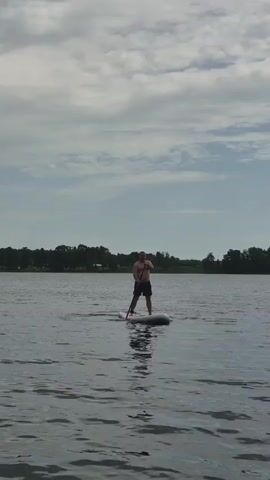 A man stands on a paddleboard in the middle of a body of water. He is wearing dark shorts and is shirtless. He holds a paddle and is in the process of paddling. The water is calm with a few ripples. In the background, there is a treeline with houses visible. The sky is cloudy with patches of blue. The video is shot from a low angle, looking up at the man and the sky.