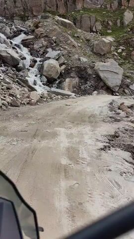 This is a first-person perspective video of a dirt road winding through a rocky, mountainous terrain. A waterfall is visible on the left side of the road, cascading down a steep, rocky slope. Large boulders are scattered throughout the scene, some on the roadside and others in the path. The road surface is uneven and appears to be made of dirt and gravel, with tire tracks visible. The camera is positioned low, possibly from a vehicle like a motorcycle, with parts of the vehicle, such as a windshield and handlebars, visible in the foreground. The overall environment is rugged and appears to be a remote, natural landscape.