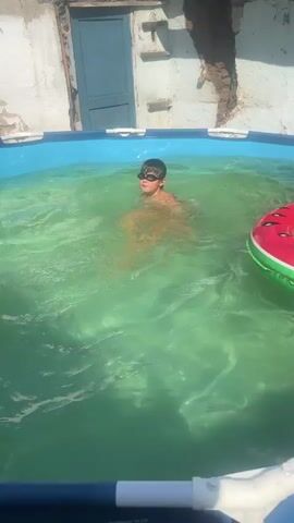 The video shows a young boy wearing goggles, standing in a shallow inflatable swimming pool filled with green water. He is in an outdoor setting, with a weathered concrete building and a blue door visible in the background. The sun appears to be shining brightly, suggesting it is daytime. The boy is actively playing in the water, moving his arms and smiling, indicating a joyful and playful mood. One of his hands is slightly raised, as if gesturing or reaching out. The overall scene depicts a child enjoying a summer day by swimming and playing in a portable pool.