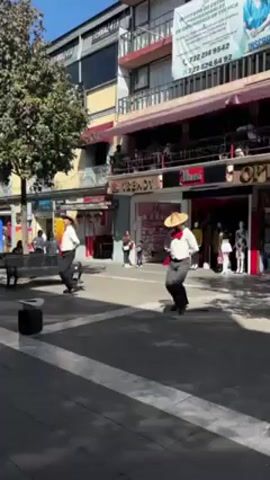 Two men in white shirts, black pants, and straw hats are tap dancing in the middle of a pedestrian street. A speaker is on the ground near them. The street is lined with shops and restaurants. People are walking and sitting on benches in the background.