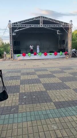 Two people are performing on an outdoor stage. The stage is covered by a metal structure with a black backdrop. There are speakers on either side of the stage. A white banner stretches across the front of the stage, decorated with red and green balloons arranged in a floral pattern. The ground in front of the stage is paved with interlocking square bricks in a checkered pattern. The sky above is a soft blue with some clouds, suggesting it is late afternoon or early evening. The performers are wearing traditional clothing. The person on the left is wearing a red and white outfit, and the person on the right is wearing a white dress. They appear to be singing or speaking.