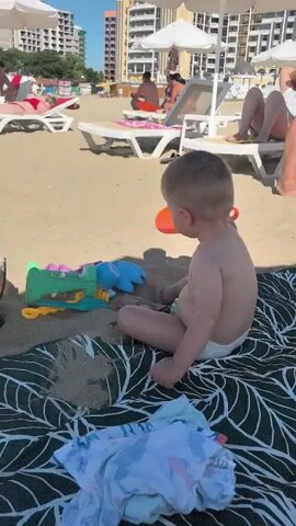 A young child wearing a diaper is sitting on a beach blanket on the sand. The child is playing with sand toys, including a shovel, a rake, and several molds. In the background, there are beach chairs, umbrellas, and people. The child is holding an orange shovel and appears to be interacting with the sand.